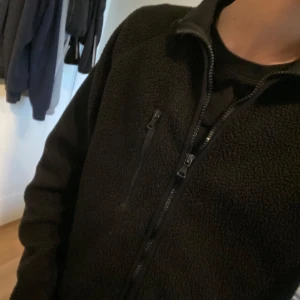 Svart fleecejacka från HM Move - Svart fleecejacka från HM Move med hel dragkedja framtill och en bröstficka med dragkedja. Jackan har hög krage och lång ärm, perfekt för lager-på-lager. Enkel och stilren design som funkar till det mesta.