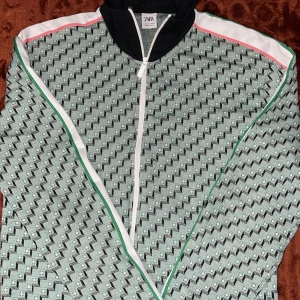 Mönstrad kofta från Zara M - Snygg hoodie från Zara i storlek M med svart huva och hel dragkedja. Jackan har ett unikt geometriskt mönster i grönt och svart, samt vita, gröna och rosa detaljer längs ärmar och axlar. Perfekt för dig som gillar sportig och trendig stil.