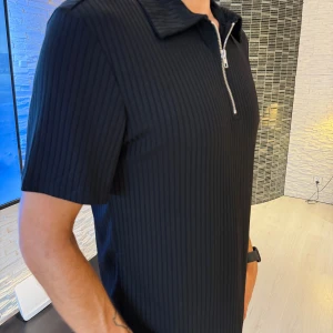 Svart ribbad t-shirt med dragkedja - Svart ribbad t-shirt med kort ärm och krage. Tröjan har en snygg silverfärgad dragkedja framtill som går ner till bröstet. Materialet känns mjukt och följsamt, perfekt för en clean och stilren look. Passar perfekt som oversize modell.