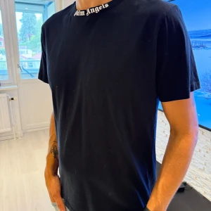 Svart Palm Angels t-shirt med tryck - Svart t-shirt från Palm Angels i mjuk bomull. Klassisk passform med korta ärmar och rund halsringning. Vit Palm Angels-logga tryckt både på kragen och stort över ryggen. Enkel men stilren streetwear-vibe. Passar perfekt som oversize modell.
