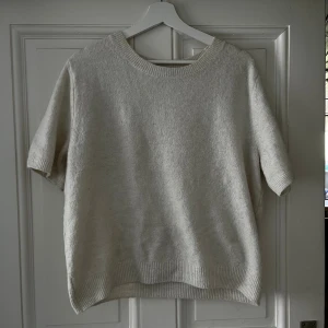 Vit/beige stickad kortärmad tröja H&M - Supermjuk och stilren vit/beige stickad tröja från H&M med korta ärmar och rund halsringning. Perfekt för lager-på-lager och funkar året runt. Ribbstickade detaljer vid hals, ärmslut och nederkant ger en clean look.