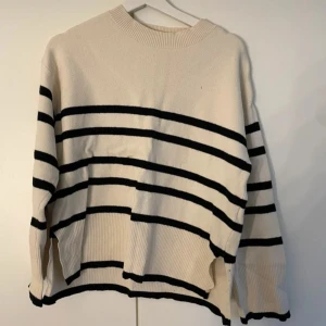 Randig stickad tröja i beige och svart - Snygg stickad tröja i beige med svarta horisontella ränder. Tröjan har en rund halsringning, långa ärmar och ribbade muddar vid ärmslut och nederkant. Perfekt för dig som gillar en avslappnad och trendig stil.