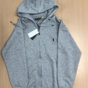Grå hoodie från Polo Ralph Lauren - Snygg grå hoodie från Polo Ralph Lauren med dragkedja framtill och klassisk logga broderad på bröstet. Tröjan har huva med snörning och två fickor framtill. Perfekt för en chill och stilren look.