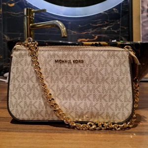 Beige axelväska från Michael Kors - Snygg beige axelväska från Michael Kors med guldfärgad kedja och dragkedja. Väskan har ett diskret mönster med MK-logga, detaljer i brunt skinn och stilren form. Perfekt storlek för det viktigaste och med lyxig känsla tack vare gulddetaljerna. Som ny och självklart äkta. 