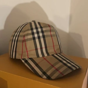 Burberry rutig keps i beige och svart - Snygg keps från Burberry med klassiskt rutigt mönster i beige, svart, vitt och röda detaljer. Kepsen har böjd skärm och justerbar rem baktill. Tillverkad i bomull, perfekt för dig som vill ha en stilren och ikonisk accessoar.