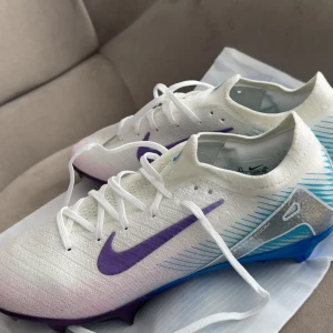 Nike Mercurial fotbollsskor elite vit/lila - Nike Mercurial fotbollsskor i vitt med lila swoosh och blå detaljer. Skorna har snörning, platt sula med dobbar och är tillverkade i syntetmaterial. Perfekta för dig som vill sticka ut på planen med stilrena och sportiga färger.