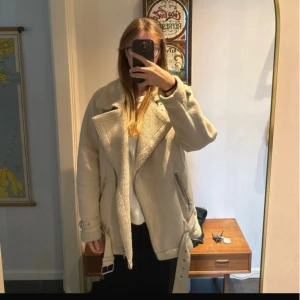 Beige mockajacka med bälte och dragkedjor - Säljer en beige mockajacka med bred krage och mjukt foder. Jackan har silverfärgade dragkedjor, bälte i midjan och detaljer i syntetmaterial. Modellen är loose fit och har långa ärmar. Perfekt för kalla dagar och har en cool, oversized look.