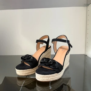 Svarta platåsandaler med kedjedetalj - Snygga svarta sandaler med hög platåsula i rep och öppen tå. Skorna har en bred rem framtill med svart kedjedetalj och justerbart spänne runt ankeln. Perfekta för sommaren och ger en trendig look till alla outfits.