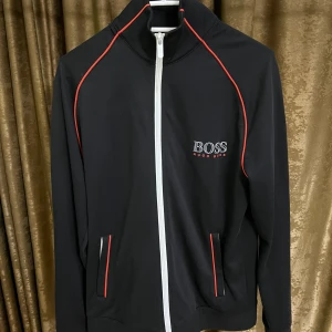 Kofta från Hugo Boss - Svart track kofta från Hugo Boss med vita dragkedjor och röda detaljer längs axlar och fickor. Jackan har hög krage, två fickor framtill och BOSS-logga på bröstet. Perfekt för en sportig och stilren look.