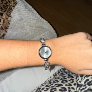 Silverfärgad armbandsklocka med stenar - Snygg armbandsklocka i silverfärg med rund urtavla och armband dekorerat med glittrande stenar. Klockan har en stilren och elegant design som verkligen sticker ut på handleden. Perfekt accessoar för dig som gillar lite extra bling.