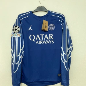 PSG långärmad blå fotbollströja - Snygg blå PSG fotbollströja med långa ärmar, vita detaljer och vingmönster på ärmarna. Officiella loggor från Jordan och Paris Saint-Germain, samt Qatar Airways tryck på bröstet. UEFA Foundation-märke på ärmen och D. Doué 14 på ryggen. Tillverkad i lätt och ventilerande material.