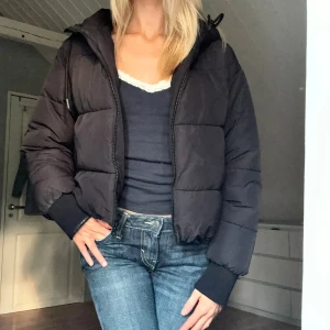 Svart pufferjacka från Monki - Svart pufferjacka med huva och dragkedja framtill. Jackan har en croppad passform med muddar vid ärmslut. Perfekt för kalla dagar och enkel att styla med det mesta 🖤                  Den är märkt med röd tuschpenna på lappen för att jag skulle veta att de va min! Annars inga defekter.