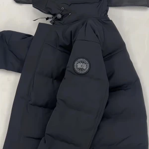 Svart pufferjacka från Canada Goose Macmillan  - Svart pufferjacka från Canada Goose MacMillan black label med huva och diskret logotyp på ärmen. Jackan har dragkedja framtill och är tillverkad i slitstark polyester med dunfyllning för extra värme. Perfekt för kalla dagar och har en clean, modern look. Fler bilder vid önskemål, pris går att diskuteras vid snabb affär 