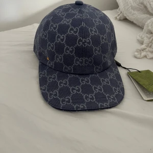 Blå keps från Gucci med GG-mönster - Snygg blå keps från Gucci med ikoniskt GG-mönster över hela kepsen. Tillverkad i bomull och har en klassisk böjd skärm. Märkets logga syns tydligt och kepsen har en exklusiv känsla, perfekt för dig som vill sticka ut.