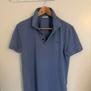 Blå pikétröja från Tiger of Sweden - Snygg blå pikétröja från Tiger of Sweden med klassisk krage och tre knappar framtill. Tröjan har korta ärmar och är tillverkad i mjuk bomull, perfekt för en stilren och avslappnad look.