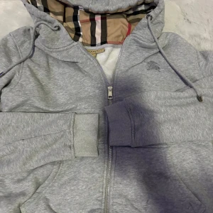 Grå hoodie från Burberry med dragkedja - Säljer en grå hoodie från Burberry i storlek L. Tröjan har dragkedja, snörning i huvan och den klassiska Burberry-rutiga insidan av huvan. Broderad logga på bröstet och mjukt foder. Perfekt för dig som vill ha en stilren och lyxig look.