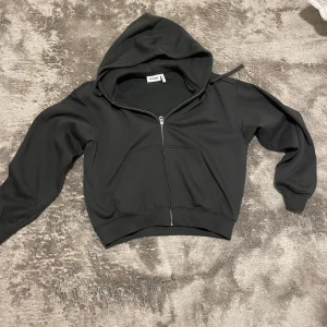 Svart hoodie med dragkedja från Weekday - Svart hoodie från Weekday i storlek XS. Tröjan har huva, hel dragkedja framtill och två stora fickor. Tillverkad i mjukt material med ribbade muddar vid ärmslut och nederkant. Perfekt för en avslappnad och stilren look.