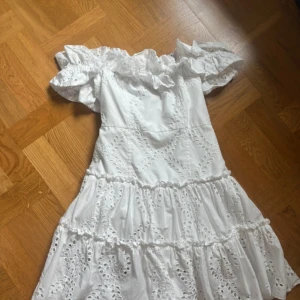 Vit offshoulder klänning från Zara - Superfin vit klänning från Zara med offshoulder och volangdetaljer. Klänningen har broderade spetsmönster och är tillverkad i bomull. Perfekt för sommarens alla tillfällen och ger en romantisk vibe med sina puffärmar och luftiga kjol.