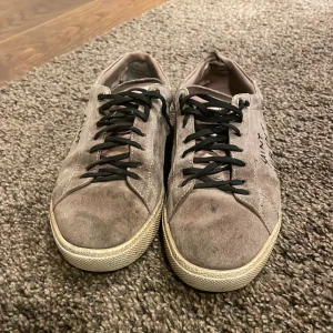 Saint Laurent sneakers i mocka - Snygga sneakers från Saint Laurent i ljusbrun mocka med svart snörning och handskriven logga på sidan. Skorna har rund tå och platt sula, perfekt för dig som gillar en stilren men ändå edgy look.