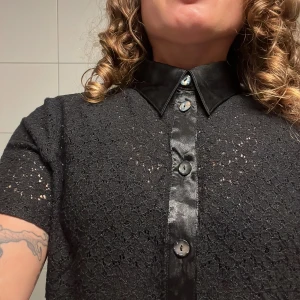 Vintage Svart spets skjorta  - Vintage Svart kortärmad skjorta från Postie Fashions med glansig krage och knappslå. Skjortan har ett genombrutet spetsmönster och pärlemorskimrande knappar. Perfekt för dig som gillar unika detaljer och en lite edgy look.