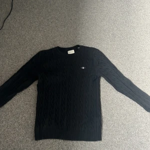 Svart kabelstickad tröja från GANT - Svart kabelstickad tröja från GANT i storlek M. Klassisk rund hals och logga på bröstet. Tröjan har långa ärmar och ribbade muddar vid hals, ärmslut och nederkant. Perfekt för dig som gillar stilrena plagg. Hör av dig vid frågor!🤝