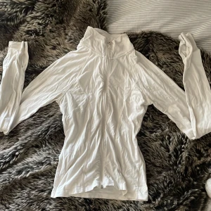 Vit ziptröja från Gina Tricot - Slimmad vit ziptröja från Gina Tricot med lång ärm och tumhål. Tröjan har hög krage och hel dragkedja framtill. Tillverkad i mjuk bomullsmix som känns skön mot huden. Perfekt för lager-på-lager och chill dagar.