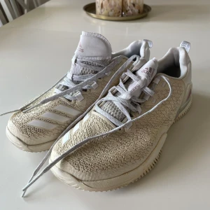 Beige Adidas sneakers med mönstrad ovandel - Snygga sneakers från Adidas i beige och vitt med coolt strukturerad ovandel och klassiska tre ränder på sidan. Skorna har grå snörning, vit sula och detaljer i syntetmaterial. Perfekta för dig som gillar sportig stil och vill sticka ut lite extra.