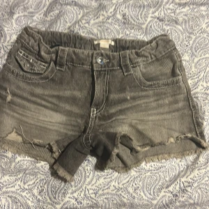 Grå jeansshorts med broderad ficka - Snygga grå jeansshorts från Gina Tricot i storlek 164. De har fransig kant nedtill, slitna detaljer och en cool bakficka med blå och vit broderad paisleymönster. Klassisk femficksmodell med knapp och dragkedja framtill.
