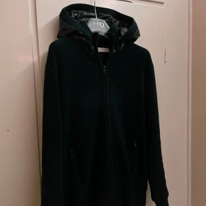 Sayless cardigan  - Mörkblå stickad jacka från sayless med dragkedja och två fickor framtill. Jackan har en glansig, quiltad huva i syntetmaterial som ger en sportig vibe. Perfekt för dig som gillar en clean och stilren look med lite edge.