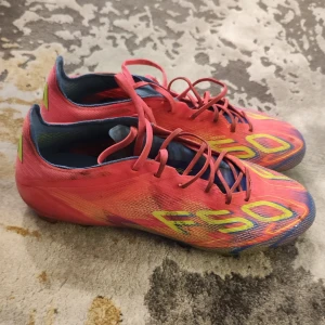 Adidas F50 rosa fotbollsskor - Säljer ett par Adidas F50 fotbollsskor i en riktigt snygg rosa färg med gula och blå detaljer. Skorna har snörning och är tillverkade i lätt syntetmaterial för bästa känsla på planen. Perfekta för dig som vill sticka ut och ha grym kontroll på bollen.