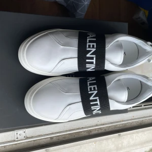 Valentino Open vita sneakers strl 44 - Säljer ett par vita Valentino Open sneakers i skinn med bred svart elastisk rem över foten där loggan står tryckt i vitt. Snygg minimalistisk design med rund tå och platt sula. Perfekta för dig som gillar exklusiva och stilrena sneakers. Om du funderar över något skriv så svarar jag, priset är inte hugget i sten vid snabb affär 😊