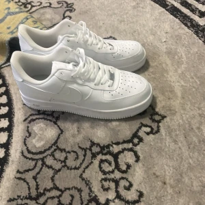 Nike Air Force 1 helvita sneakers - Klassiska Nike Air Force 1 sneakers i helvitt skinn med perforerad tå, rund tå och platt sula. Snygg och tidlös modell med snörning och ikonisk Swoosh-logga på sidorna. Perfekta för dig som vill ha en clean och stilren look. Helt OANVÄNDBAR 