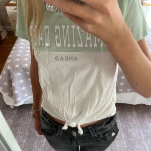Mintgrön och vit t-shirt från H&M - T-shirt från H&M i mintgrönt och vitt med trycket 'AMAZING DAYS AHEAD' framtill. Modellen är kortärmad och har en knytning nertill för en trendig look. Tillverkad i mjuk bomull, perfekt för en avslappnad stil. Typ aldrig använd så super skick passar mig som har  xs-s. Hör av er om frågor!💕
