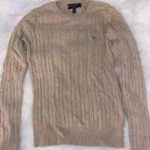 Beige kabelstickad tröja från Gant - Kabelstickad beige tröja från Gant i stretchig bomull. Klassisk rund halsringning och diskret broderad logga på bröstet. Perfekt för lager-på-lager och enkel att matcha med jeans eller kjol.