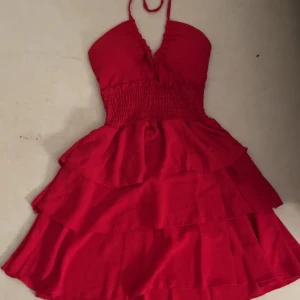 Röd volangklänning med halterneck - Supersöt röd kortklänning med halterneck och smockad midja. Klänningen har flera volanglager i kjolen och knytning i nacken. Perfekt för dig som vill sticka ut med en färgstark look. Materialet känns lätt och luftigt. Insydd bh skydd. Aldrig använd!