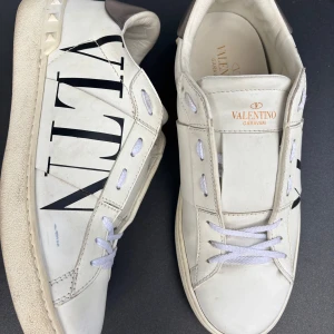 Valentino Garavani vita sneakers - Snygga vita sneakers från Valentino Garavani med svart VLTN-logga på sidan och coola nitar runt hälen. Skorna har silvriga detaljer baktill och klassisk snörning. Tillverkade i skinn för en lyxig känsla och stilren look.