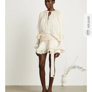 Champagnefärgad omlottklänning med volang - Superfin kort klänning i champagnefärgat, lätt och glansigt material. Den har långa, vida ärmar, omlottknytning i midjan och volangig kjol. Snygg knytning vid halsen och flowig siluett som ger en lyxig känsla.Ny pris 1500kr