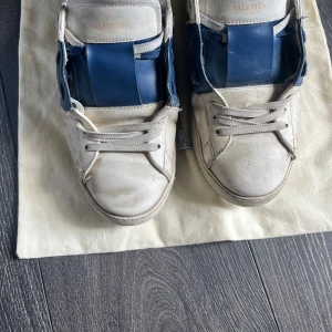 Valentino Garavani sneakers blå/vit - Snygga sneakers från Valentino Garavani i vitt skinn med bred blå läderdetalj över vristen. Klassisk rund tå, platt sula och coola nitar på hälen. Vit snörning och diskret logga på plösen. Perfekta för dig som gillar exklusiva och trendiga skor.