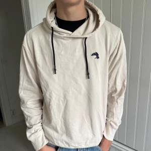 Hoodie  - Snygg beige hoodie från The Black Label Collection med svart broderad fågel på bröstet och trycket 'Fly Above The Black Sky' på ryggen. Tröjan har svarta snören i huvan och är tillverkad i mjuk bomull. Hör av dig vid frågor! 