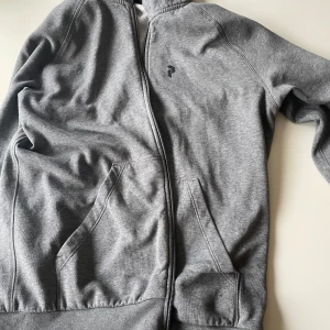 Grå hoodie från Peak Performance - Säljer en grå hoodie från Peak Performance med dragkedja, huva och två stora fickor framtill. Tröjan har diskret logga på bröstet och är tillverkad i mjuk bomullsblandning. Perfekt för chill dagar eller när du vill ha en clean och enkel stil.