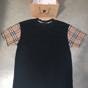 Burberry t-shirt  - Svart t-shirt från Burberry med klassiska beige-rutiga korta ärmar. T-shirten har en rak passform och är tillverkad i mjuk bomull. Ikoniskt rutmönster på ärmarna ger en lyxig och trendig vibe. Påse medföljer  oanvänd möts i Sthlm passar även S