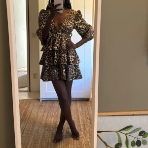 Leopardmönstrad volangklänning - Supersnygg kort klänning med leopardmönster och flera volanger. Djup v-ringning, trekvartsärmar och markerad midja. Materialet är glansigt och lätt, perfekt för att sticka ut. Dragkedja i ryggen för enkel påklädning.