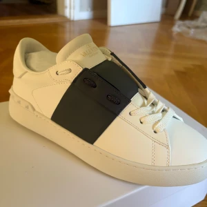 Valentino Garavani Open sneakers vit/marinblå - Säljer ett par Valentino Garavani Open sneakers i vitt skinn med bred svart läderpanel över snörningen. Klassisk rund tå, platt sula och vita snören. Snygg och stilren design med subtila nitar på hälen. Kommer med originalkartong, dustbag och tags.