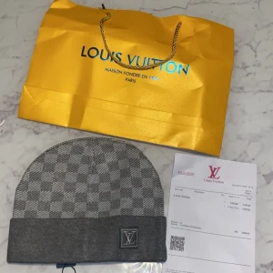 Grå rutig mössa från Louis Vuitton - Exklusiv grå mössa från Louis Vuitton med klassiskt rutigt mönster och vikt kant. Mössan har en diskret LV-logga framtill och är tillverkad i mjukt material som passar perfekt för kyliga dagar. Snygg accessoar för dig som gillar lyxiga detaljer.