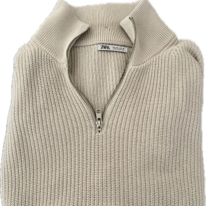 Beige stickad half zip-tröja från Zara - Beige stickad tröja från Zara med hög krage och half zip-dragkedja. Tröjan har långärmade ribbade muddar och är gjord i ett grovstickat material som ger en chill och stilren vibe. Perfekt för lager-på-lager under höst och vinter.