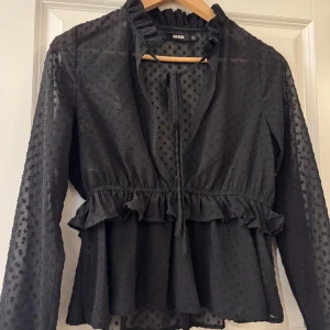 Svart prickig blus från Bik Bok - Supersöt svart blus från Bik Bok med prickigt mönster, volangdetaljer i midjan och knytning vid halsen. Blusen har lång ärm och lätt transparent tyg som ger en luftig känsla. Perfekt för dig som gillar romantiska och trendiga plagg.