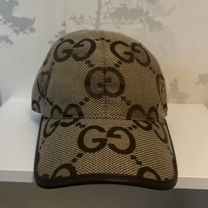 Gucci keps med GG-mönster - Snygg keps från Gucci i beige och brunt med det klassiska GG-logomönstret över hela kepsen. Justerbar rem bak i brunt skinn och detaljer i samma färg. Tillverkad i canvas med exklusiv känsla, perfekt för dig som vill sticka ut med en ikonisk accessoar. Pris ej hugget !