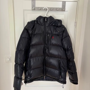 Svart pufferjacka Polo Ralph Lauren - Svart pufferjacka från Polo Ralph Lauren med huva, dragkedja och flera fickor. Jackan har en röd broderad logga på bröstet och är tillverkad i glansig polyester. Perfekt för kalla dagar och har en riktigt schysst streetstil. Orderbekräftelse finns!