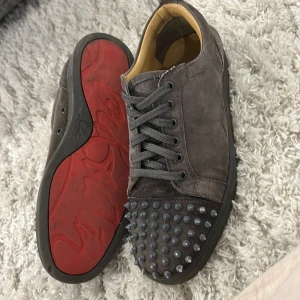 Gråa Louboutin med nitar - Säljer ett par gråa sneakers i mocka från Christian Louboutin med ikoniska röda sulor. Skorna har snörning och coola svarta nitar över tån för en edgy vibe. Insidan är beige och sulan är svart. Perfekta för dig som vill sticka ut.