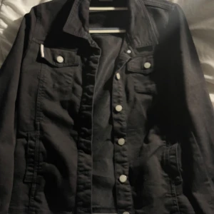 Svart jeansjacka med silverknappar - Stilren svart jeansjacka med klassisk krage och snygga silverfärgade knappar framtill. Jackan har två bröstfickor med lock och knappar samt två sidofickor. Perfekt för dig som vill ha en tidlös och cool look.